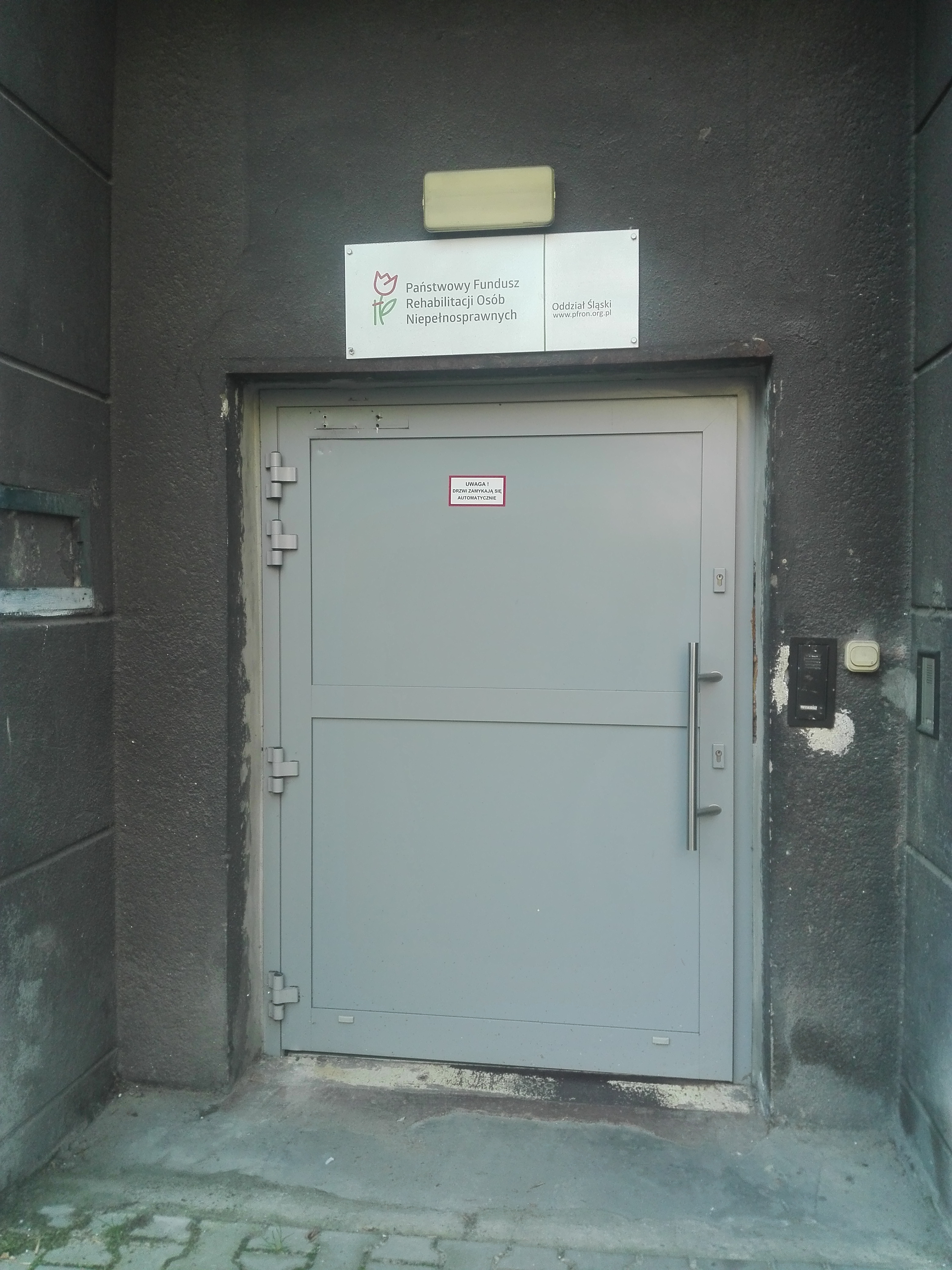 Szerokie jasnoszare drzwi. Nad drzwiami tablica z logo i napisem „Państwowy Fundusz Rehabilitacji Osób Niepełnosprawnych”. Po prawej stronie od drzwi dzwonek oraz domofon. Na drzwiach tabliczka z napisem „uwaga, drzwi zamykają się automatycznie”
