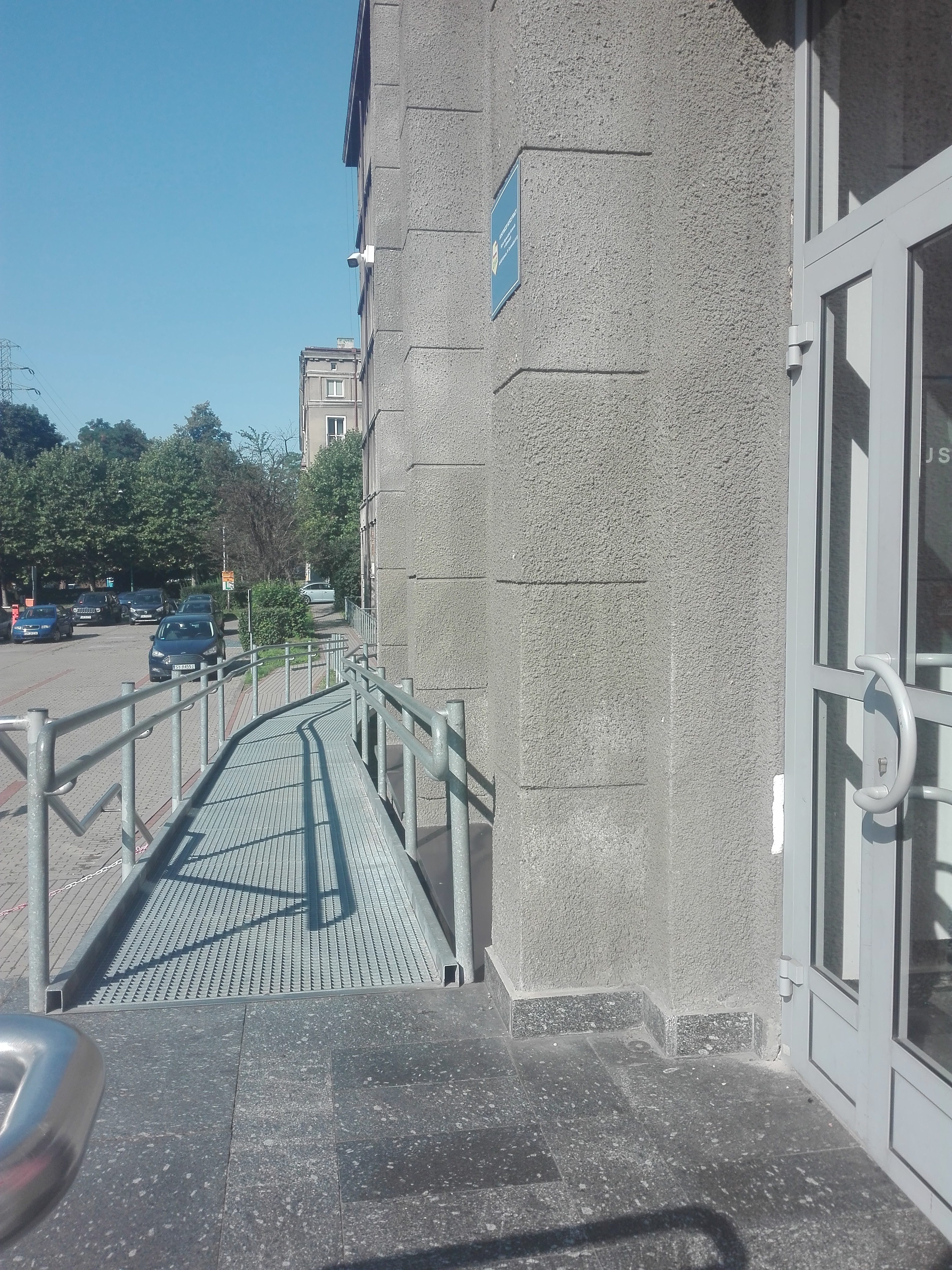 Widok z wejścia do budynku na pochylnię dla wózków i jej przebieg wzdłuż budynku z 2 zakrętami. W tle widać parking z zaparkowanymi samochodami. 