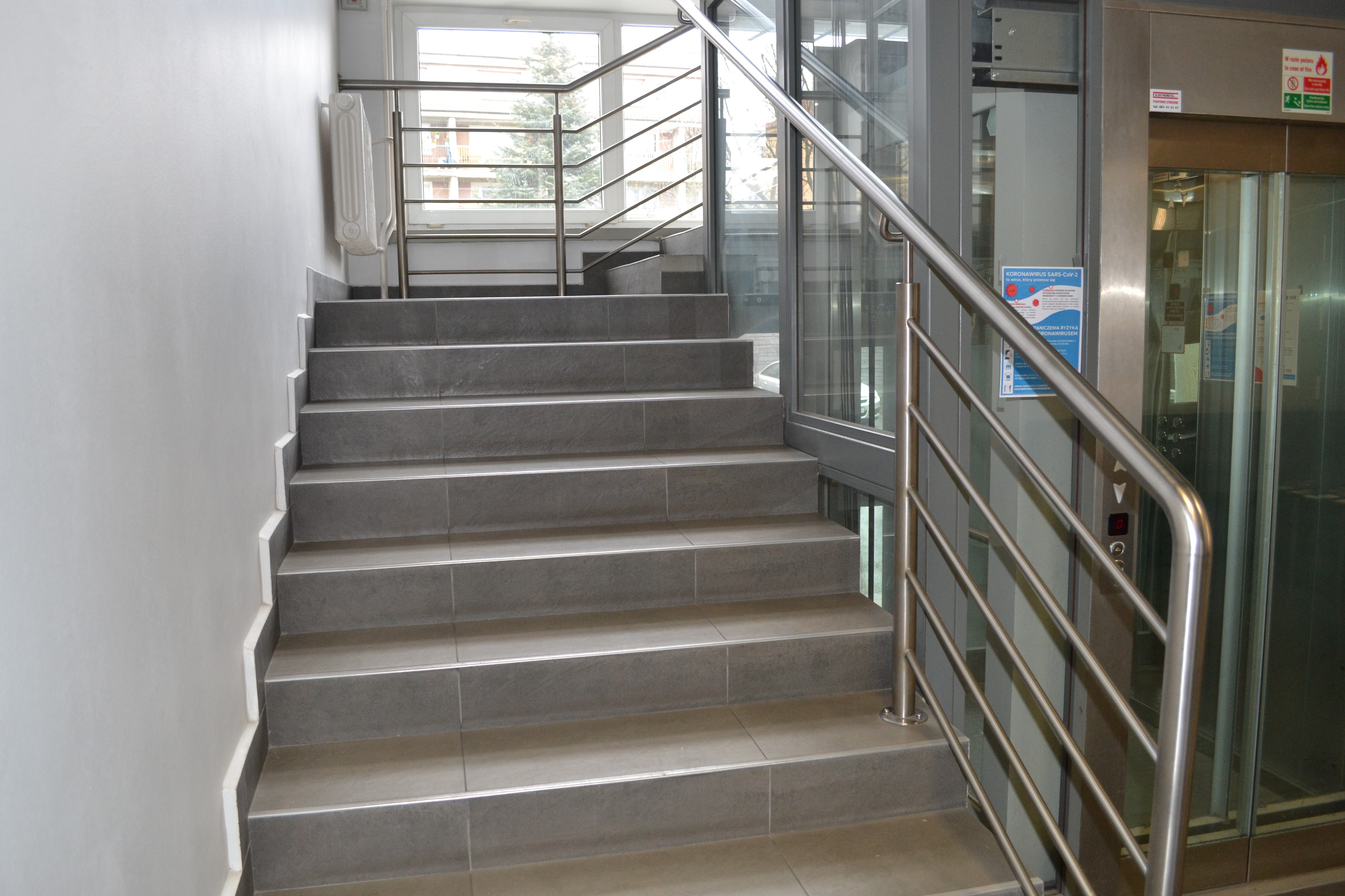 Klatka schodowa z jasnoszarymi schodami, otaczającymi szyb windy. Po prawej stronie przeszklone drzwi do kabiny.