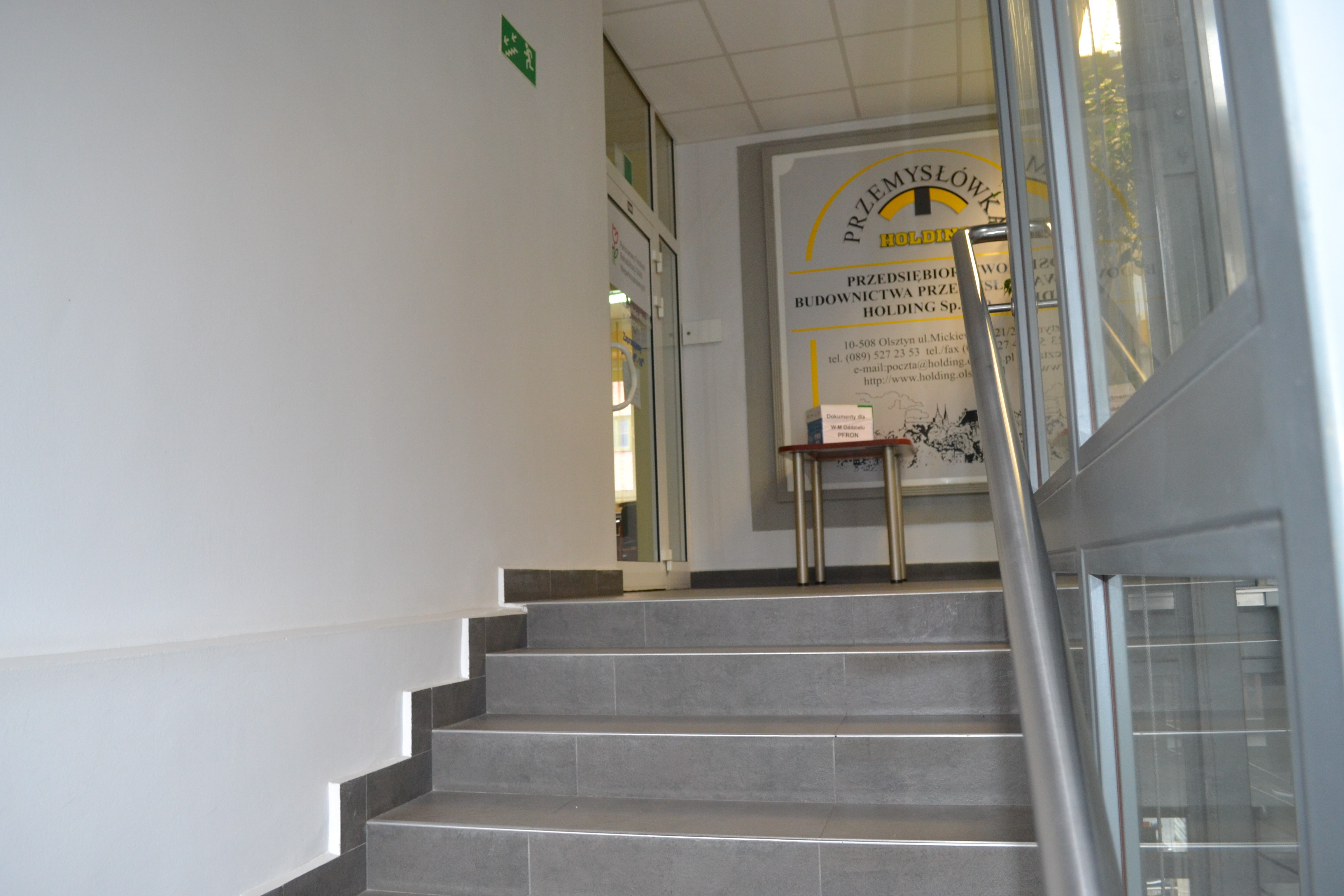 Widok na schody prowadzące do Oddziału. Jasnoszare schody z poręczami. Po prawej schodó1) szyb windy.