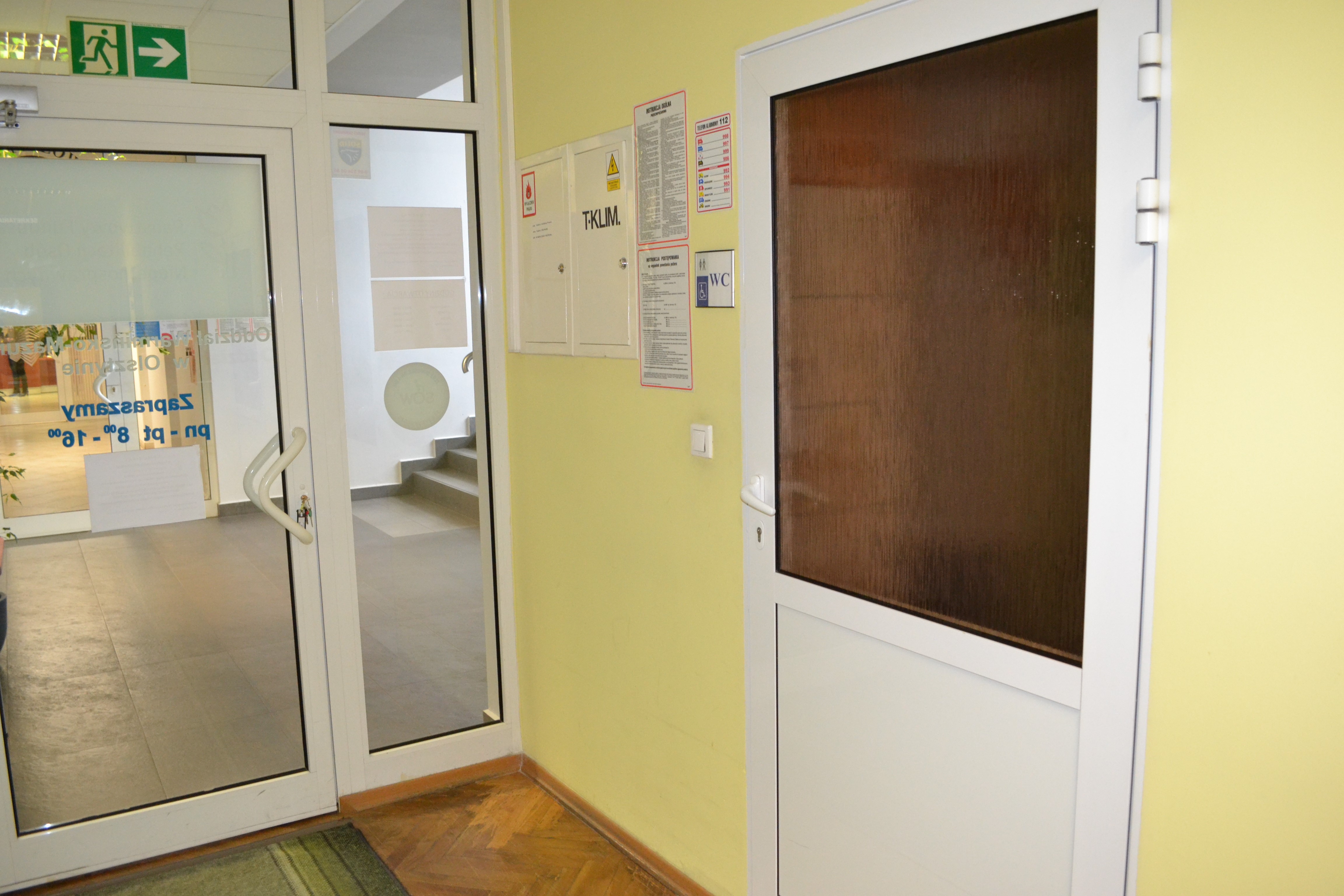 Widok na wejście do toalety i wejście do Oddziału PFRON od strony korytarza Oddziału. Na ścianie po prawej stronie widoczne skrzynki elektryczne oraz instrukcje przeciwpożarowe.