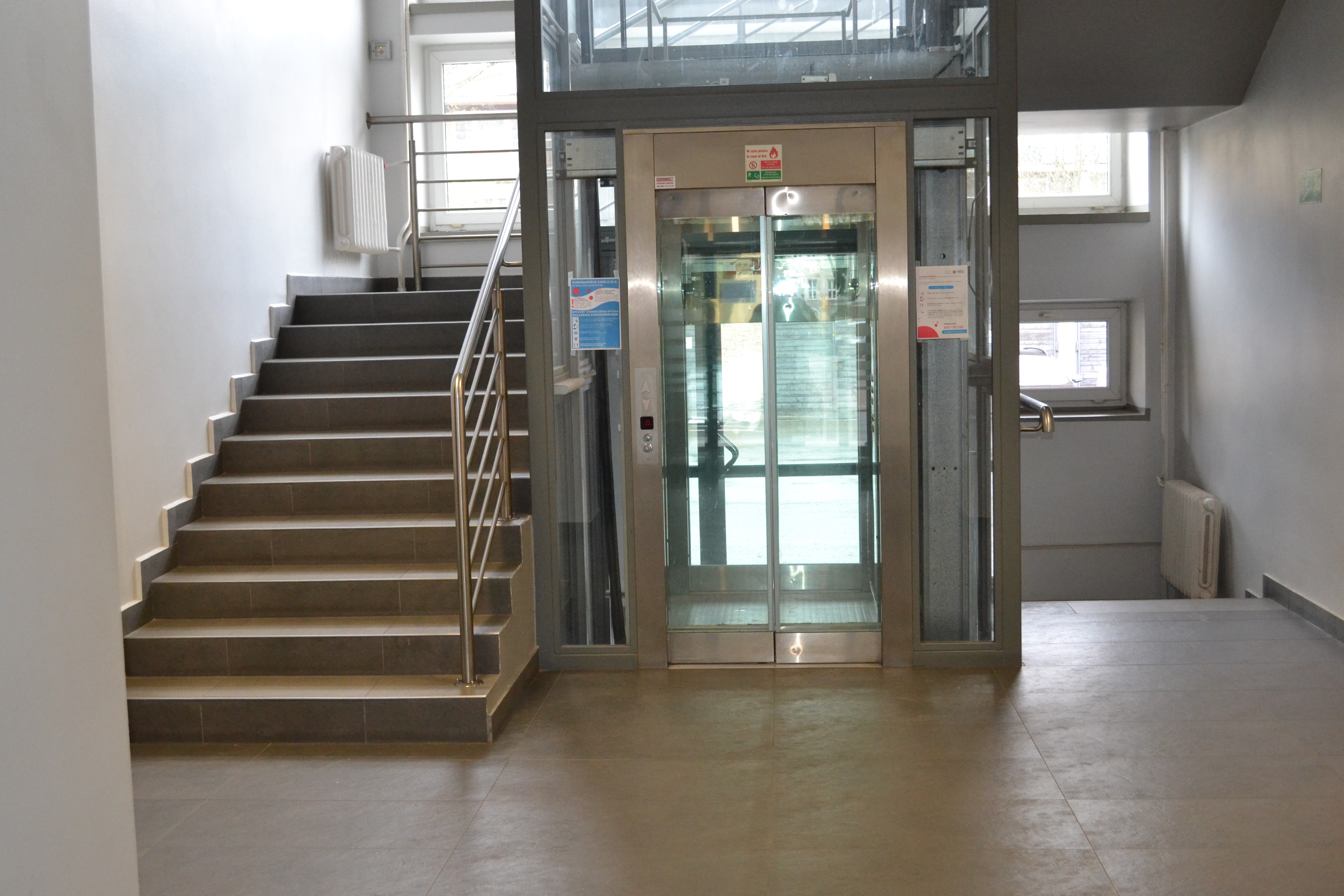 Wejście do windy wewnątrz budynku. Szerokie, przeszklone drzwi prowadzą do przestronnej, przeszklonej kabiny. Szyb windy otoczony jest klatką schodową