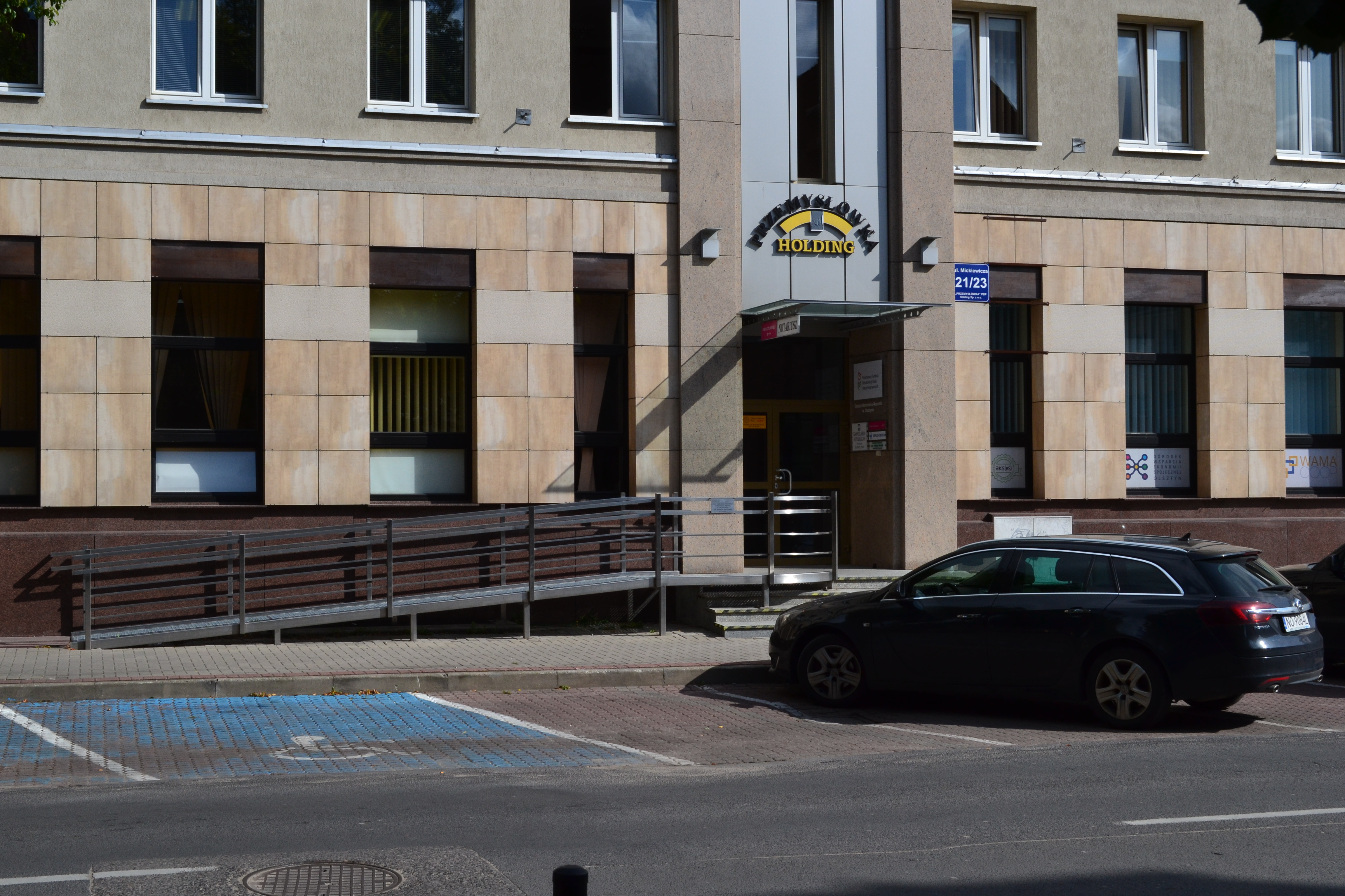 W tle budynek biurowy, obłożony kamieniem z pochylnią dla wózków. Nad wejściem napis żółtymi literami „Przemysłówka Holding”. Przed budynkiem zaparkowany samochód, a po lewej stronie do samochodu widoczne oznakowane miejsce parkingowe dla osoby niepełnosprawnej