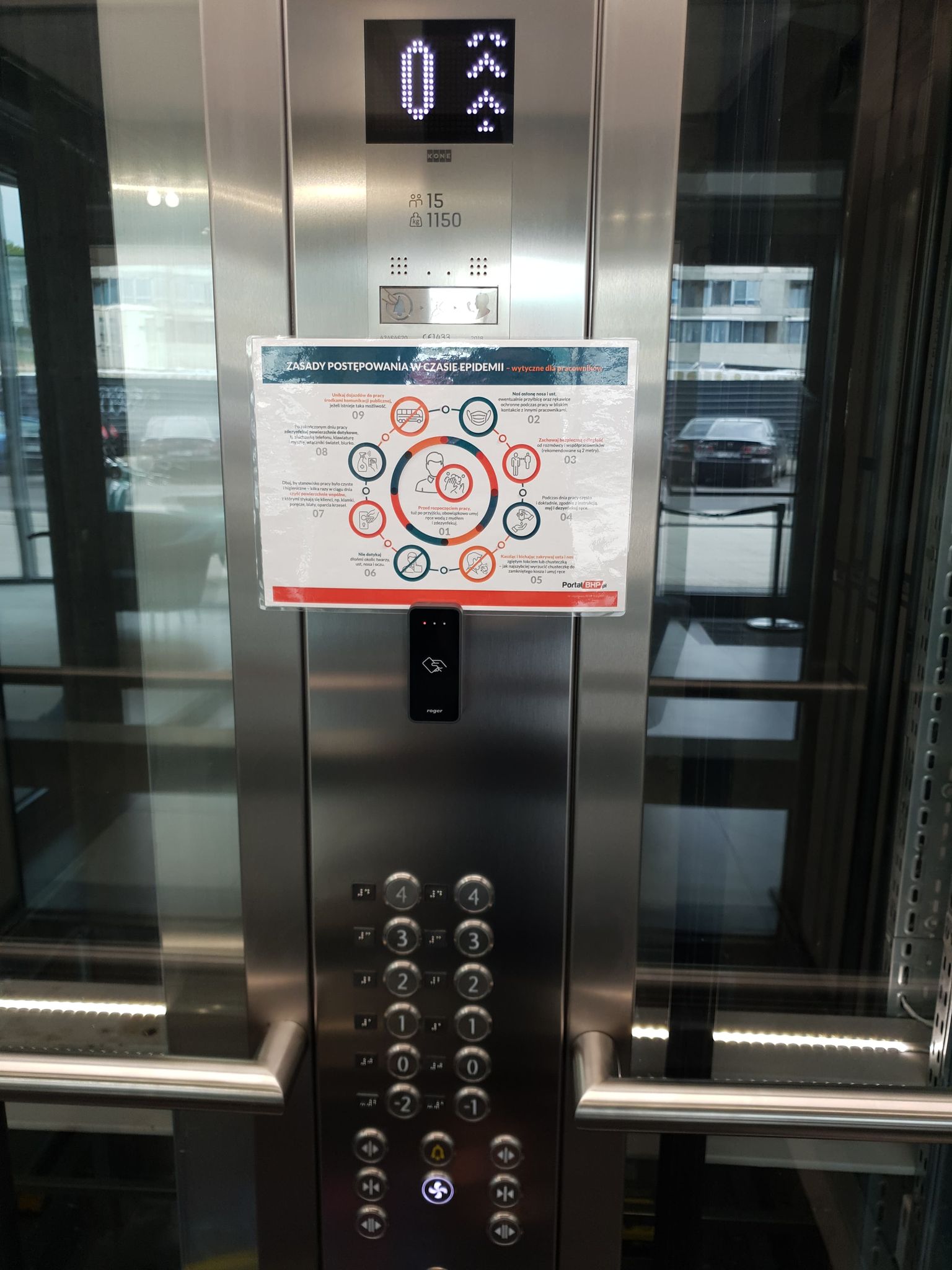 Panel windy, przyciski pięter oznaczone wypukłymi cyframi po ich prawej stronie napisy w alfabecie Braille’a. Na środku panelu naklejona kartka z informacją, jak zasad przestrzegania higieny w pandemii.