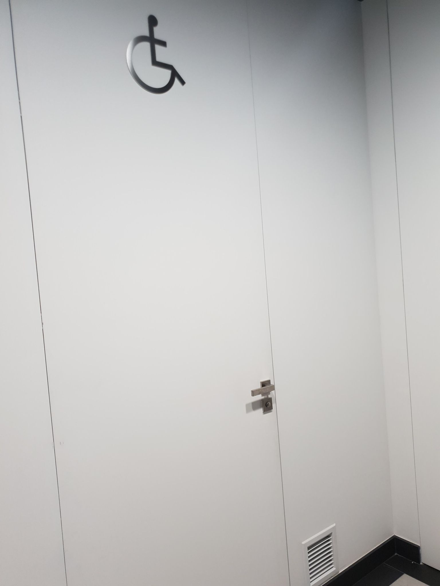 Drzwi wejściowe do toalety dla niepełnosprawnych koloru białego z dużym piktogramem osoby niepełnosprawnej. 