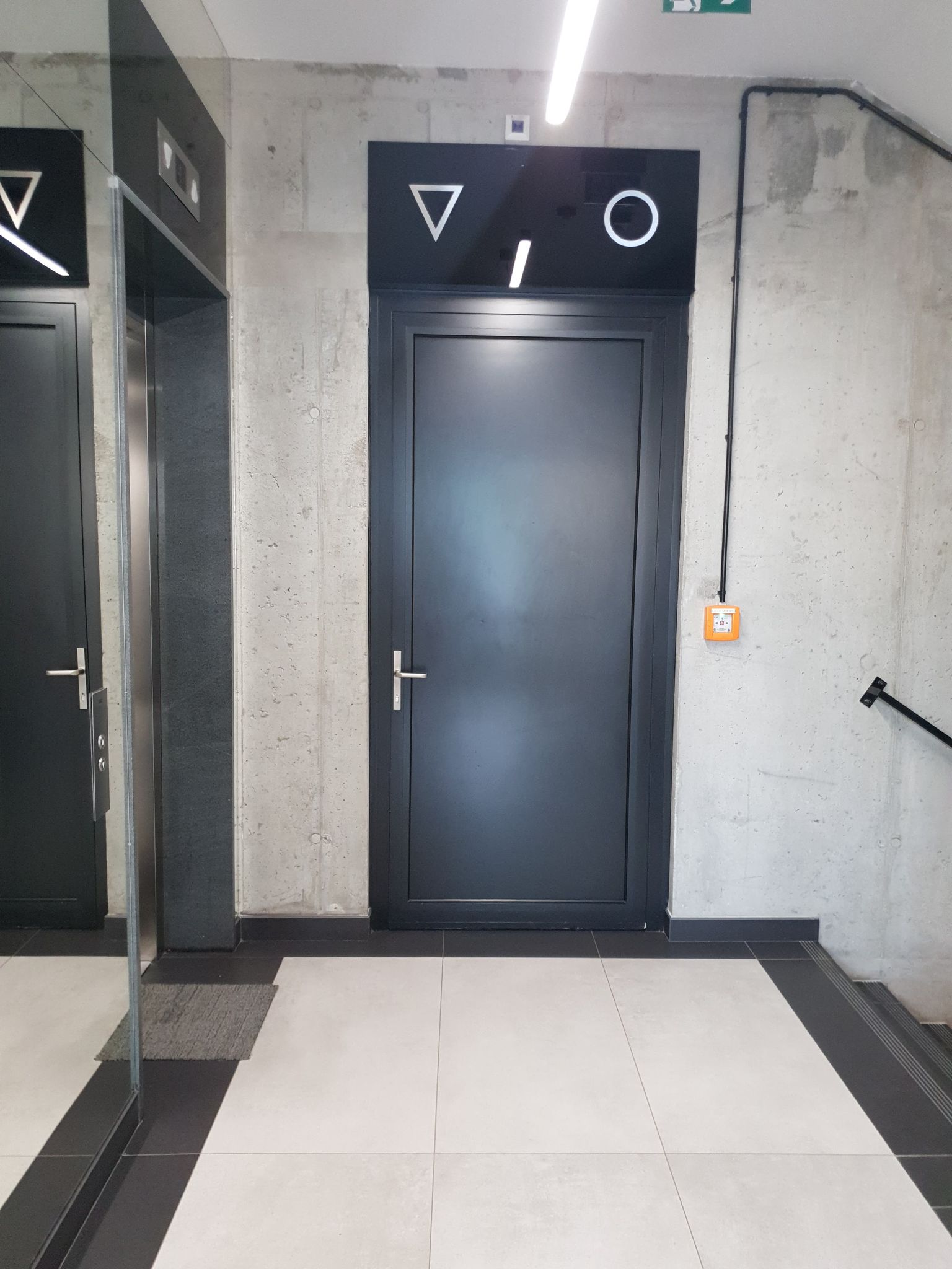 Na tle szarej ściany czarne drzwi, nad nimi oznaczenie piktogramami toalety damskiej i męskiej. Po lewej stronie od drzwi do toalety, widoczne drzwi szybu windy 