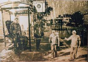 Pokaż zdjęcie: zdjęcie nieostre w odcieniach brązu, przedstawia przystanek autobusowy i 4 mężczyzn oraz w tle nadjeżdżający autobus