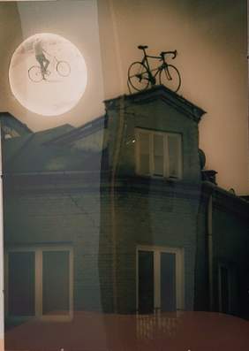 Pokaż zdjęcie: zdjęcie domu i rowerem na dachu, a nad nim księżyc w którym jest widoczny mężczyzna z rowerem