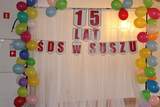 NA zdjęciu czerwony baner z napisem 15 lat ŚDS w Suszu, wokół niego kolorowe balony