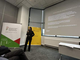 Pokaż zdjęcie: Kobieta stojąca w Sali konferencyjnej, na ekranie wyświetla się prezentacja
