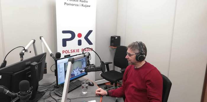 Przy stole w siedzibie Radia PiK siedzi pracownik PFRON Michał Bohuszko, w słuchawkach na uszach, przed nim stoi komputer, a w tle baner Radia PiK